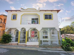 A two-story building with a white facade, yellow trim, and barred windows. - Cozy Apartment Rental in Puerto Plata, Dominican Republic (Puerto Plata)