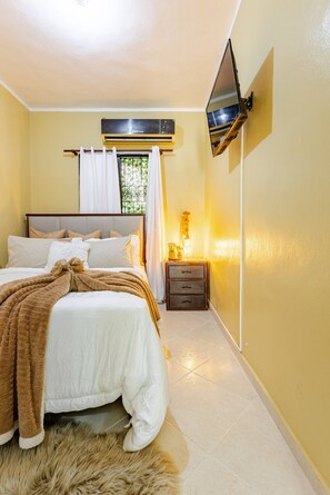 A hotel room with a bed, a TV mounted on the wall, a bedside table, and a window with curtains. - Cozy Apartment Rental in Puerto Plata, Dominican Republic (Puerto Plata)