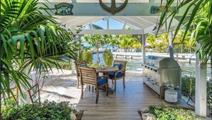 Een overdekt terras met een barbecue, houten stoelen en uitzicht op een zwembad en palmbomen.