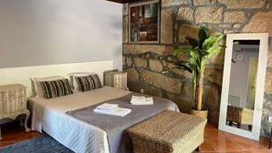 A bedroom with a stone wall, a bed with pillows, a bedside table, a plant, and a mirror.
