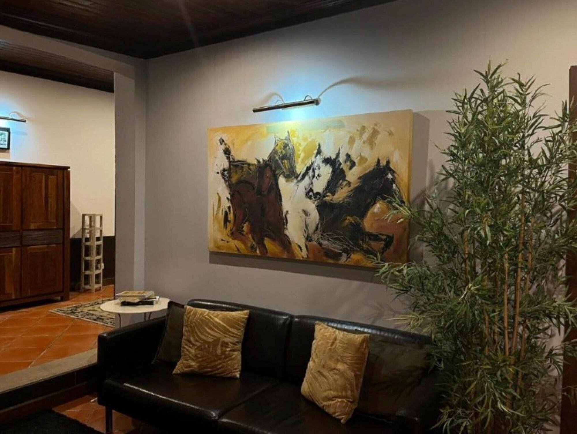 A living room with a black leather sofa, two cushions, a large abstract painting, a tall green plant, and a wooden cabinet.