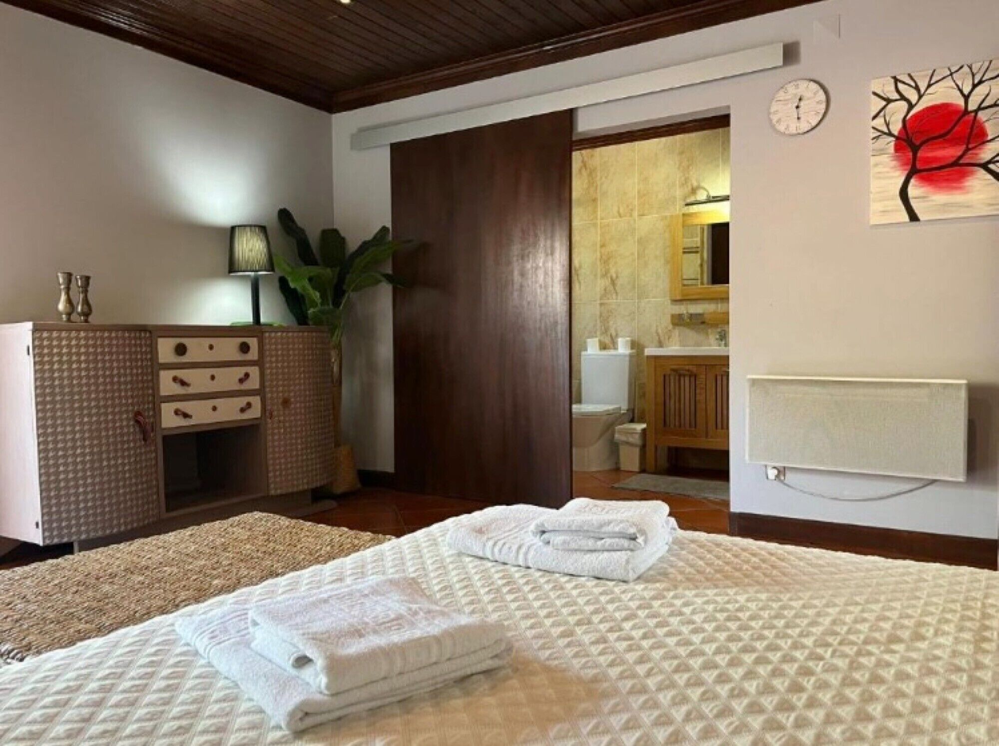A hotel room with a bed, a dresser, a plant, a painting, and a bathroom visible through an open door.