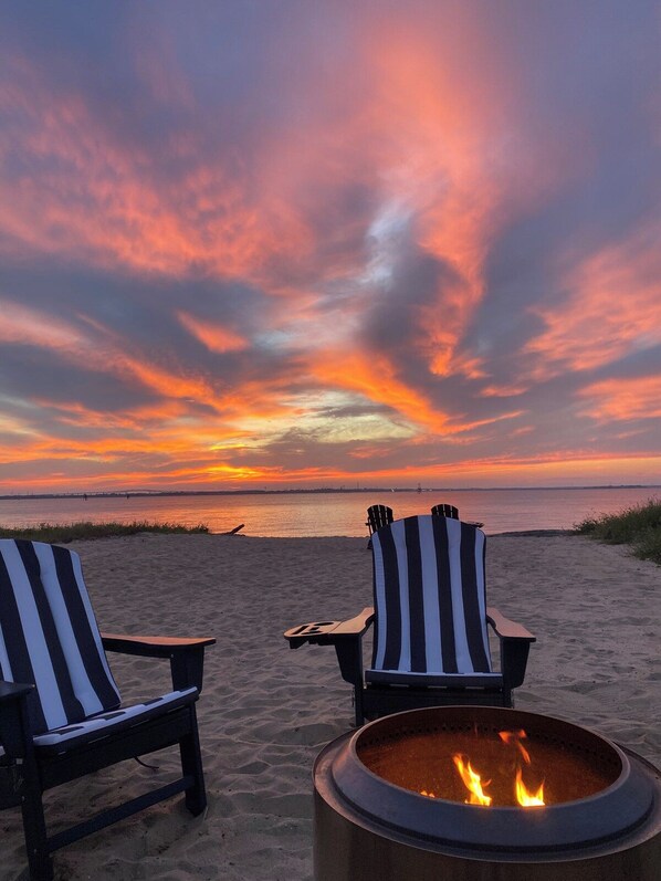 Two striped beach chairs facing a fire pit on a sandy beach with a sunset in the background.