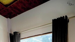 A room with a wooden table and two chairs, a window with curtains, and a view of greenery outside. - Ayu Hotels  (Ukhimath)