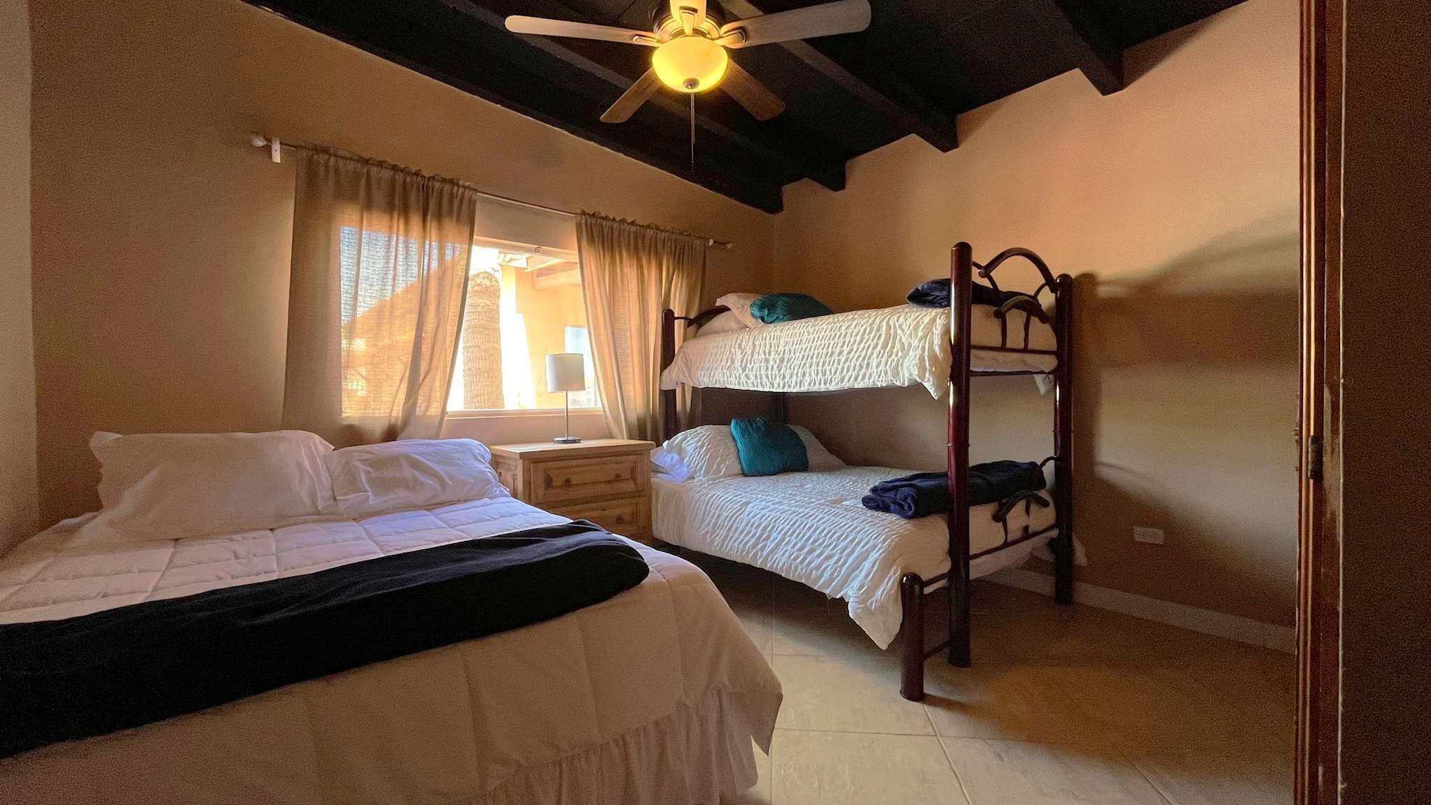 A room with a bunk bed, a single bed, a nightstand, a ceiling fan, and a window with curtains.