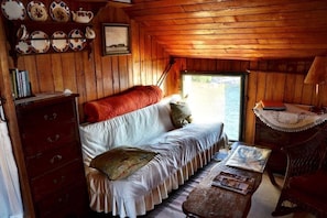 Une pièce confortable, aménagée comme un chalet, avec un plafond en bois, un canapé blanc recouvert d’une couverture rouge, une table en bois avec des magazines et une fenêtre donnant sur une étendue d’eau.