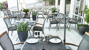Un área de comedor al aire libre con mesas preparadas para dos, sillas, plantas en macetas y una barandilla al fondo.