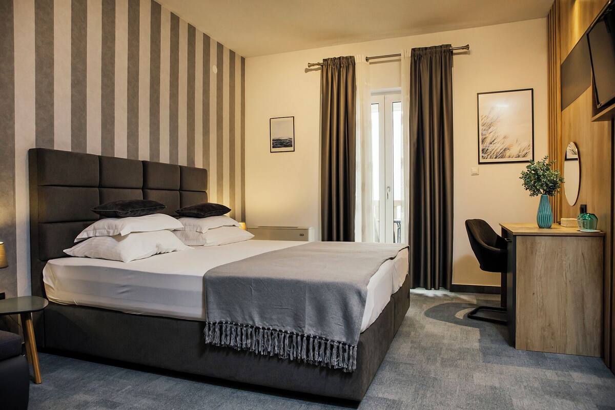 Quarto de hotel com cama, escrivaninha, cadeira, espelho e janela com cortinas.