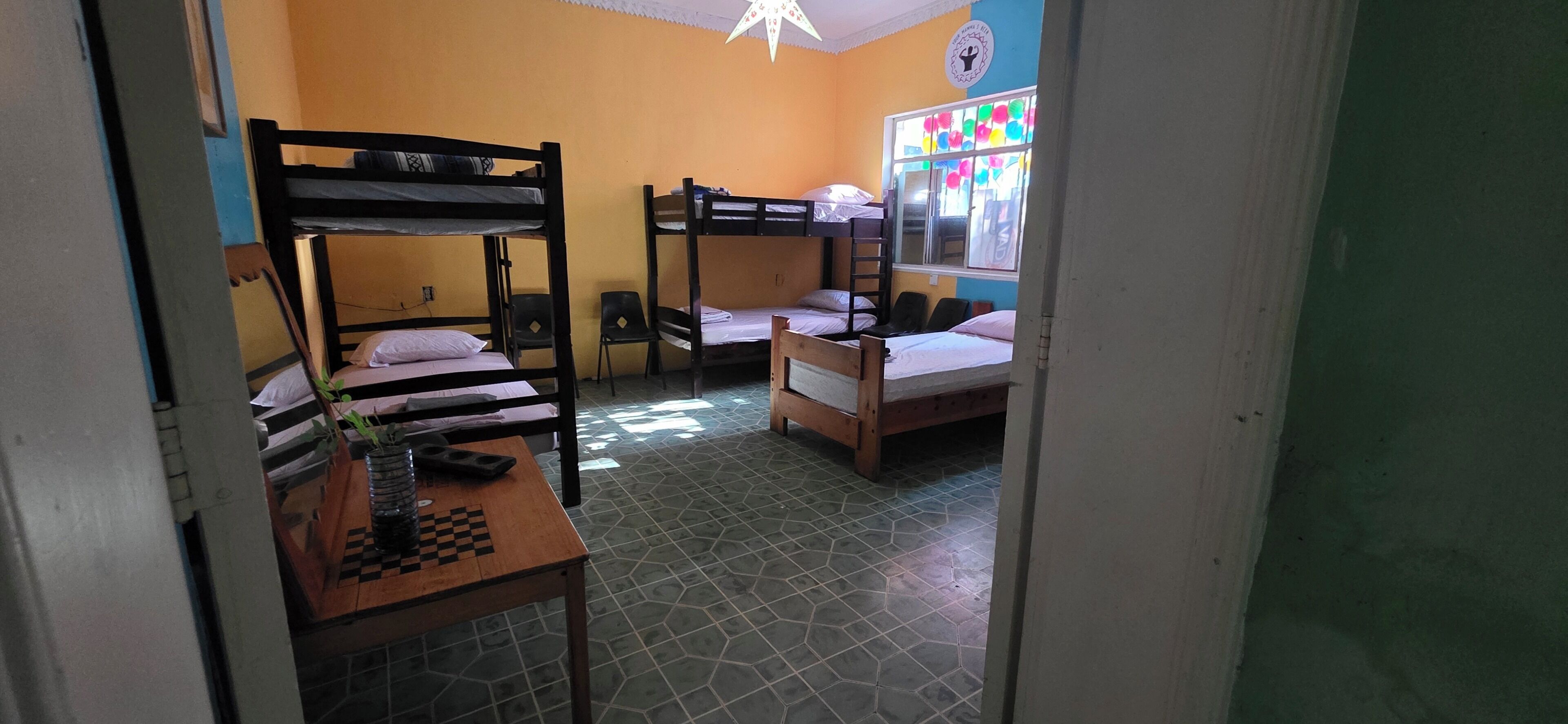 Un dormitorio con literas, una mesita con un tablero de ajedrez y una ventana con adornos coloridos.