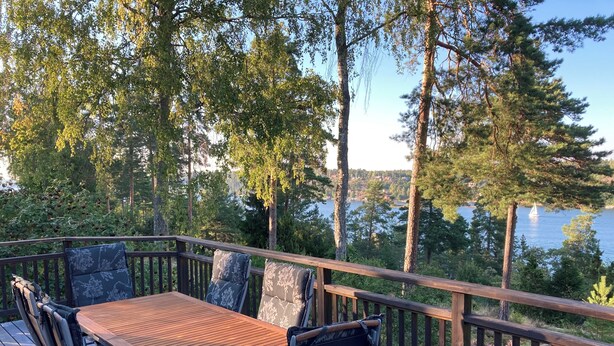 Eine Holzterrasse mit Tisch und Stühlen, mit Blick auf einen See, Bäume und ein Segelboot.