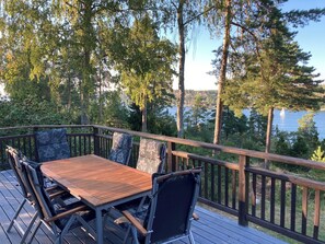 Une terrasse en bois avec une table et des chaises, donnant sur un lac bordé d’arbres et sur lequel on peut apercevoir un voilier.