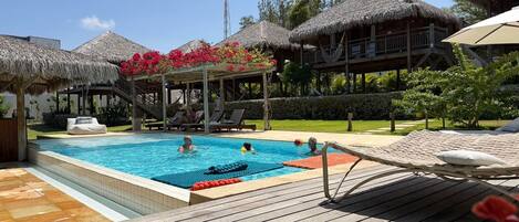 A pool area with lounge chairs, a wooden deck, and thatched-roof structures.