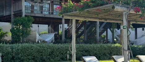 Un resort con edificios de techo de paja, una pérgola de madera con flores colgantes y un césped cuidado con sillones de descanso.