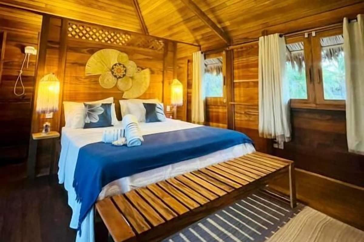 Interior de una cabaña de madera con cama, banco y lámpara colgante.