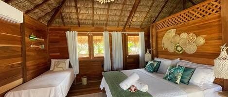 A wooden cabin with two single beds, a large bed with a green throw, and a thatched roof.