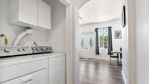 Un área de lavandería con lavadora, secadora y conducto de ventilación. Hay una puerta que da a un comedor con una mesa y sillas.