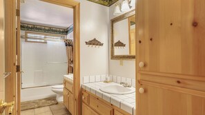 A bathroom with a bathtub, toilet, sink, and wooden cabinets. - Pinehaven Retreat by Avantstay Hot Tub + Firepit + Games + Sun Room Minutes to Slopes & Lake (Big Bear Lake)