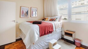 Een slaapkamer met een bed, een raam met gordijnen, een klein nachtkastje en een prullenbak.
