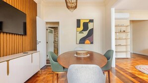 Een eethoek met een ronde houten tafel, groene beklede stoelen, een flatscreen televisie bevestigd aan een houten paneel en een modern abstract schilderij aan de muur.