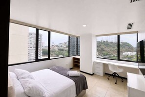 A modern hotel room with a large bed, a desk, and a chair, with a city view through large windows. - Modern 2 BR Apt on the 14th floor includes Parking (Guayaquil)