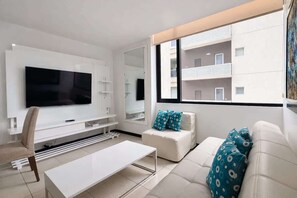 Ein modernes Wohnzimmer mit Flachbildfernseher, weißen Möbeln, einer Couch mit gemusterten Kissen und einem großen Fenster mit Blick auf ein Gebäude.