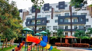 A playground with slides and climbing structures, surrounded by well-maintained grass and trees, with a modern apartment building in the background. - Elegant Apartment free parking near US Consulate (Guayaquil)