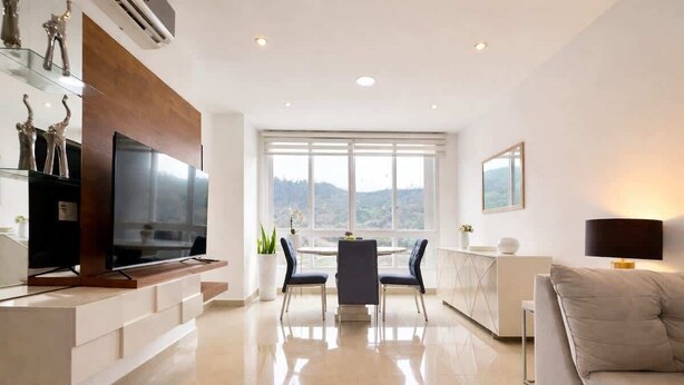 A modern living room with a flat-screen TV, a dining area with a table and chairs, a sofa, and a large window with a view. - Elegant Apartment free parking near US Consulate (Guayaquil)