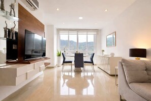 Una sala de estar moderna con televisor de pantalla plana, comedor con mesa y sillas, sofá, y un ventanal con vistas.