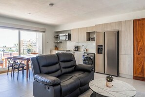 Ein stilvoller Wohnraum mit einer Couch, einem Essbereich, einer Küche mit Geräten und Blick durch große Fenster.