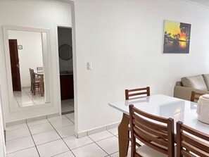 A dining area with a white table and wooden chairs, a kitchen in the background, and a painting on the wall.