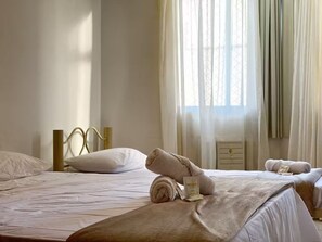A hotel room with two beds, a window with sheer curtains, and a bedside table with a rolled towel and a bottle.