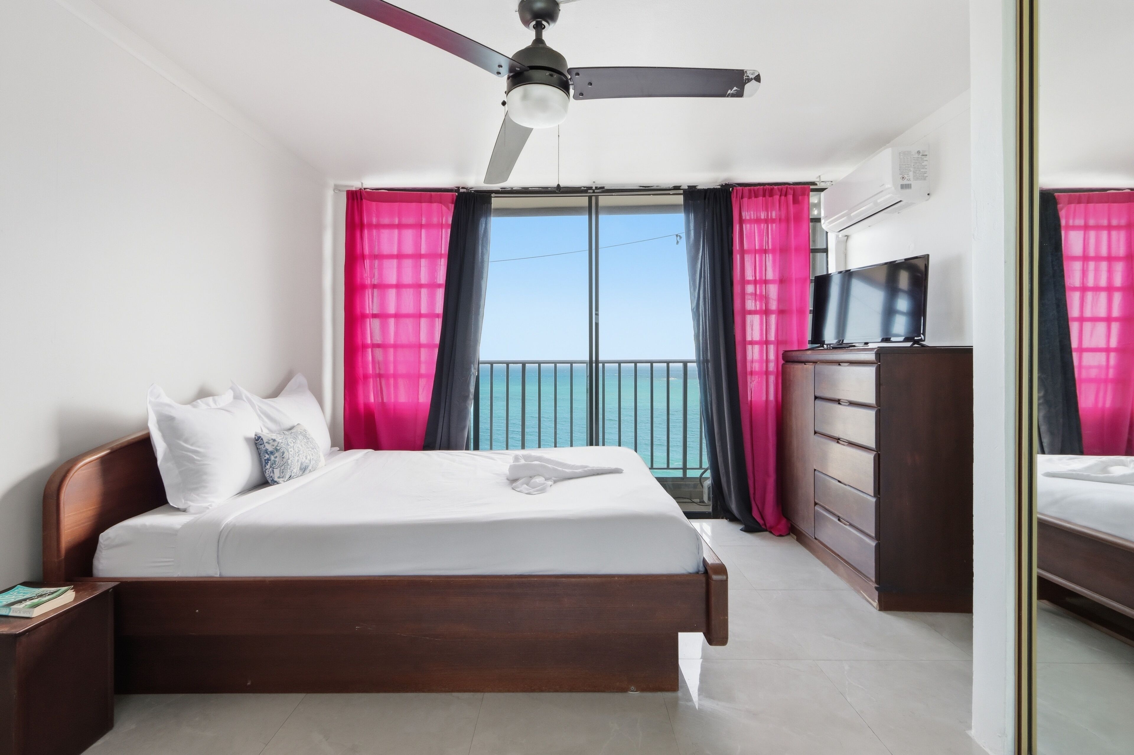Une chambre d’hôtel avec un lit, une commode, une télévision et un balcon avec des rideaux.