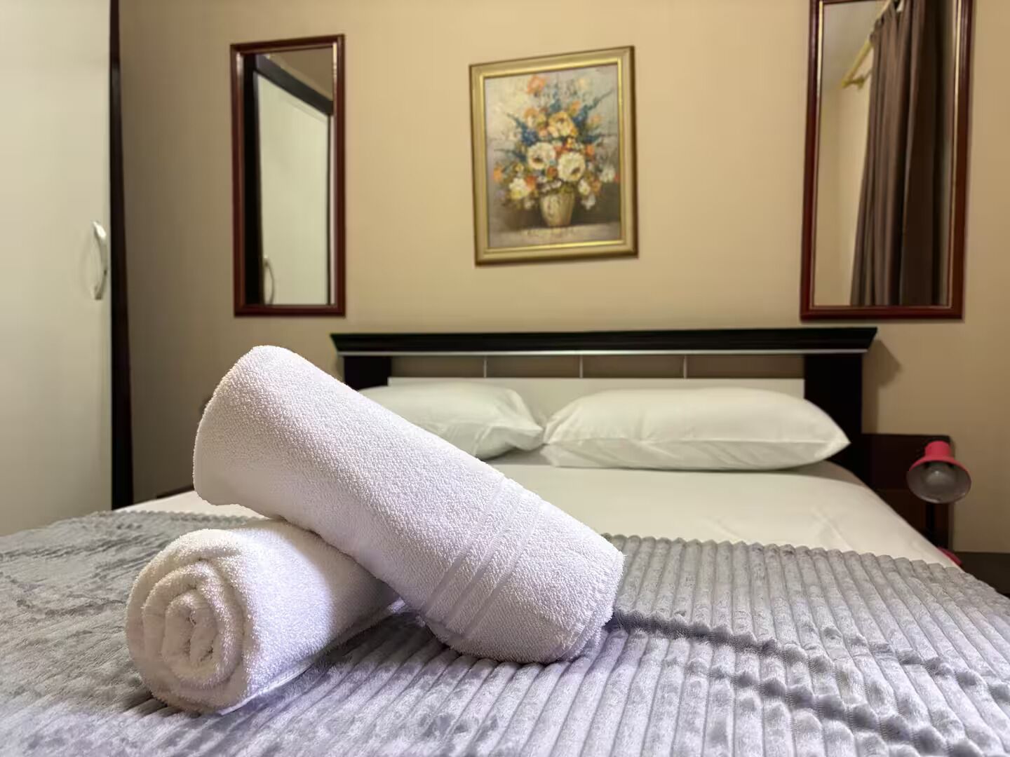 A hotel room with a neatly made bed, a painting on the wall, and a mirror above the bed.