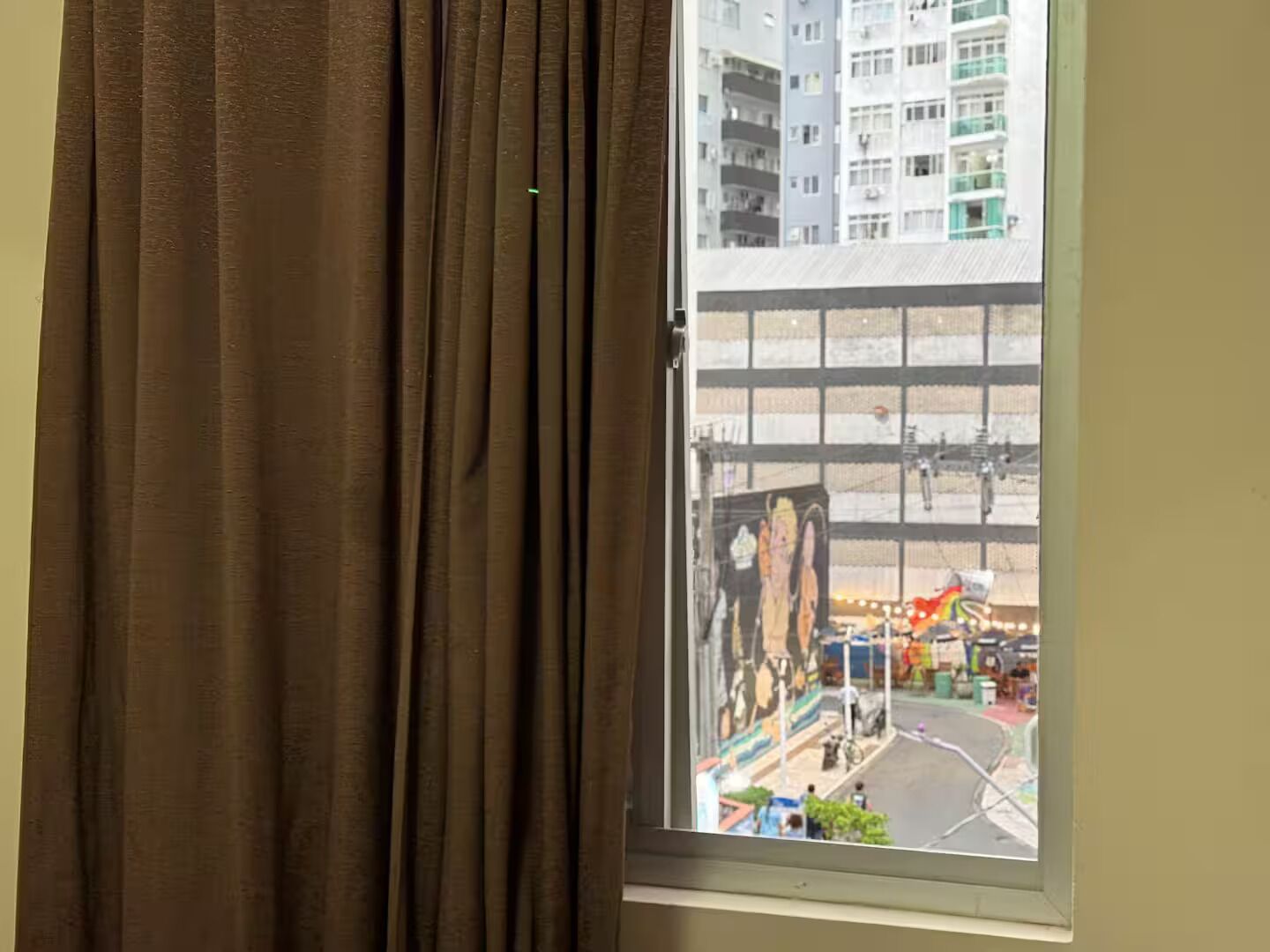 A window with brown curtains, a view of a multi-story building, and a street scene with parked cars and a mural.