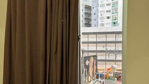 Ein Fenster mit braunen Vorhängen, Blick auf ein mehrstöckiges Gebäude sowie eine Straßenszene mit parkenden Autos und einem Wandgemälde.