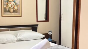 Ein ordentlich gemachtes Bett mit weißen Leinen, ein aufgerolltes weißes Handtuch, ein Nachttisch mit einer Lampe und ein Gemälde an der Wand.