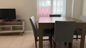 Comedor con mesa y sillas, televisor en un mueble bajo y ventana con cortinas translúcidas.