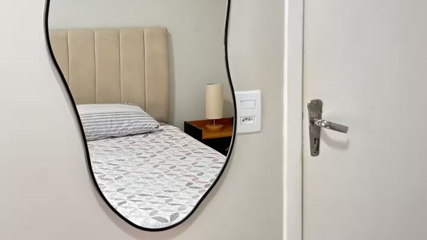 Una habitación con una cama, una mesita de noche con lámpara, un enchufe y una puerta con picaporte.