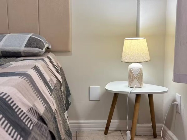 A neatly made bed with a patterned blanket, a small white table with a lamp, and a wall socket. - 2 Bedrooms, Garage and Close to the Beach (Balneário Camboriú)