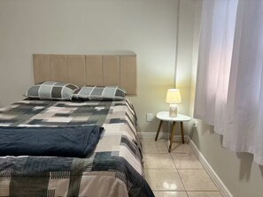 A bedroom with a bed, pillows, a bedside lamp, and a curtain. - 2 Bedrooms, Garage and Close to the Beach (Balneário Camboriú)
