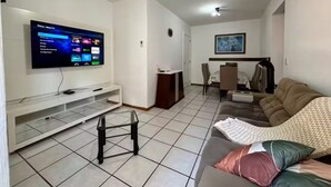 Een woonkamer met een flatscreen televisie, een bank, een salontafel en een eethoek met stoelen.