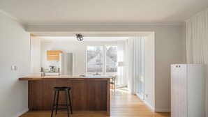 Une cuisine moderne avec un îlot central en bois, des tabourets de bar, un réfrigérateur et une grande fenêtre donnant sur la forêt.