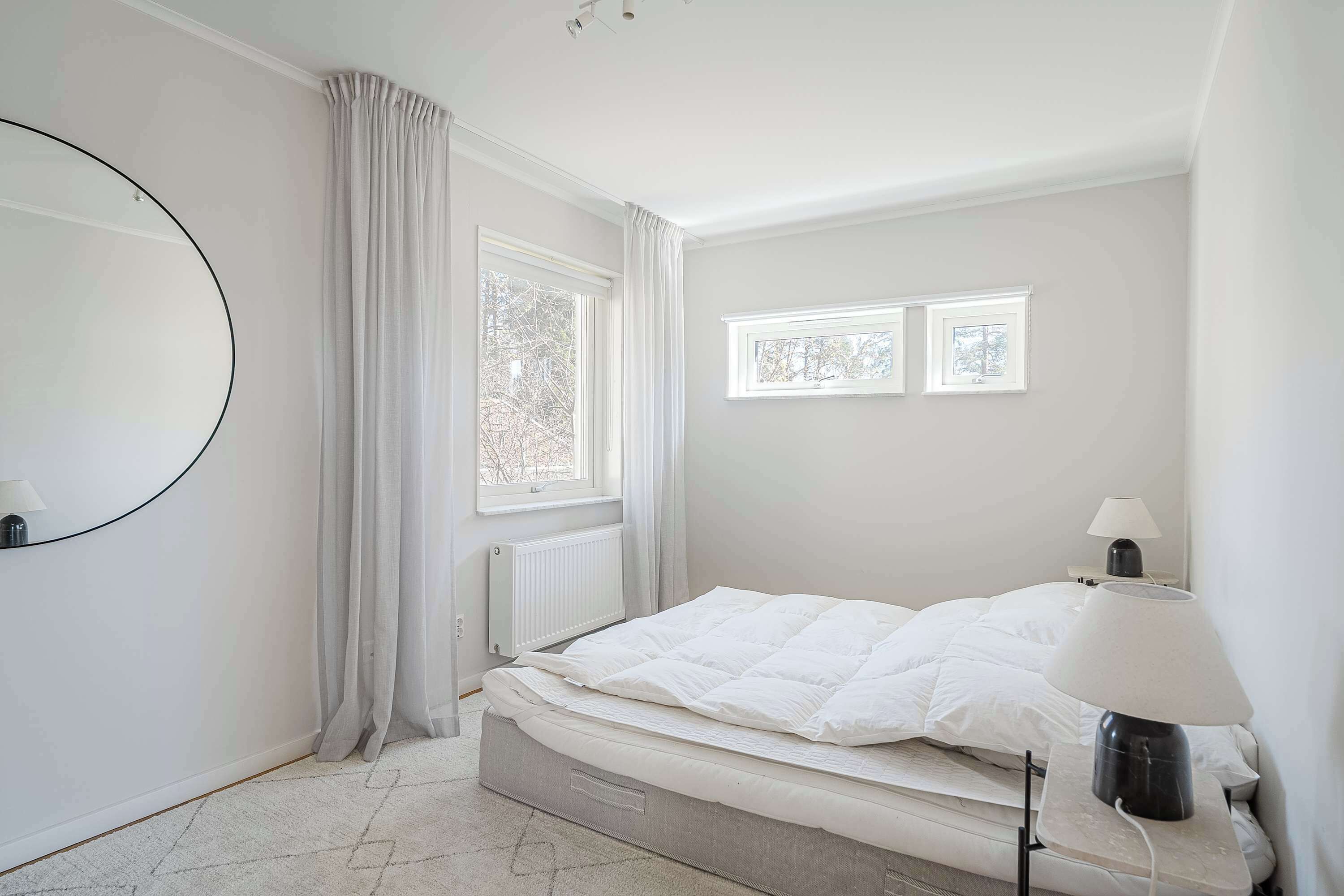 Ein Schlafzimmer mit einem Bett, zwei Fenstern, einer Heizung, einer Lampe und einem Spiegel.