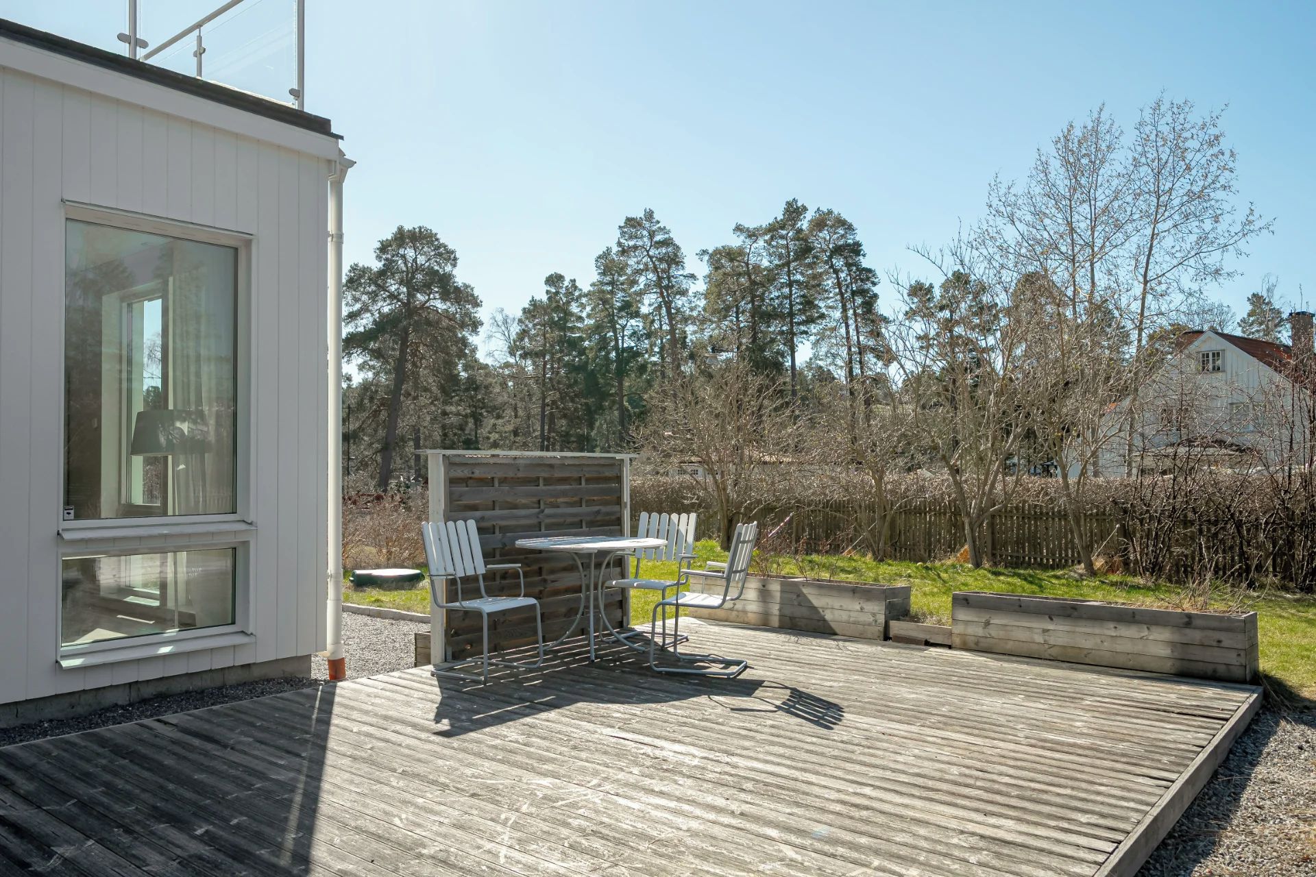 En moderne udendørs terrasse med et spisebord og stole, omgivet af et træhegn og træer.