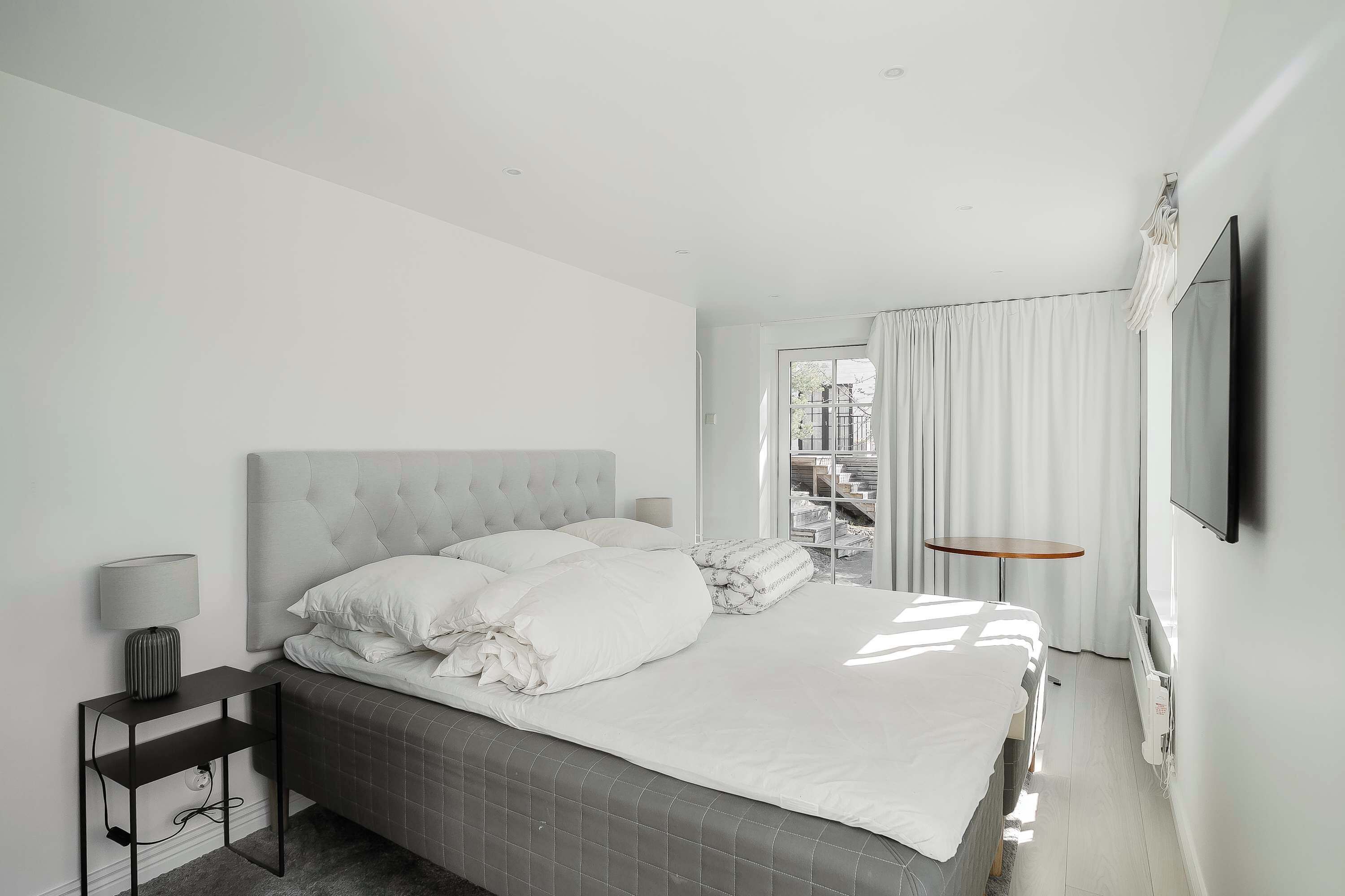 A modern hotel room with a large bed, a bedside table with a lamp, a TV mounted on the wall, and a view of the outside through the window.