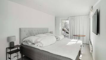 A modern hotel room with a large bed, a bedside table with a lamp, a TV mounted on the wall, and a view of the outside through the window.