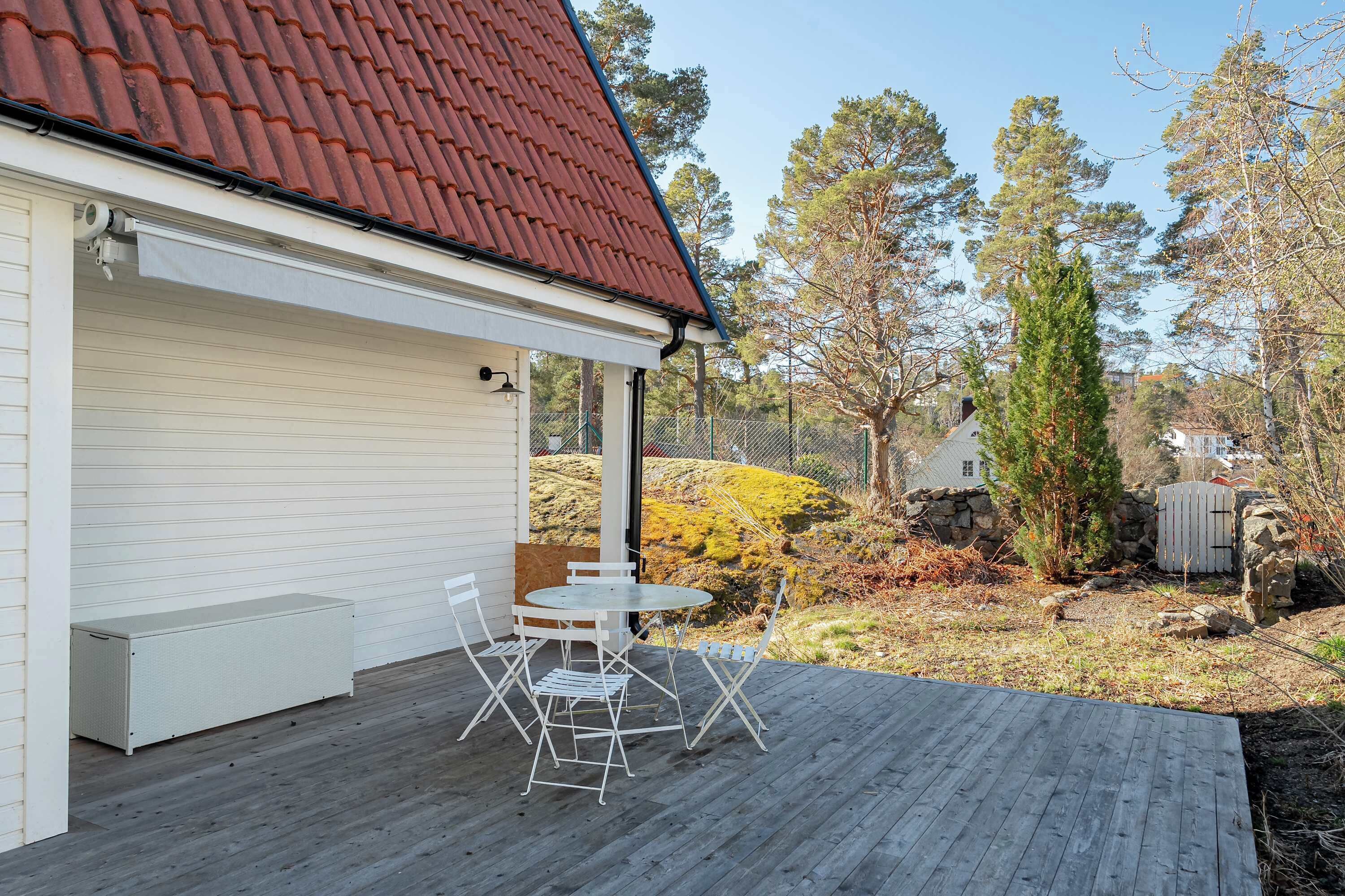 Eine überdachte Terrasse mit Tisch und Stühlen, ein weißes Gebäude mit rotem Dach und eine natürliche Landschaft mit Bäumen und Felsen.