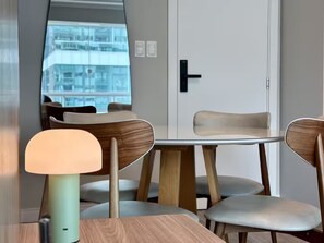 A modern room with a round table, four chairs, a lamp, and a view of a building through a large mirror. - Oceanfront near the Ferris Wheel - 1 Bedroom (Balneário Camboriú)
