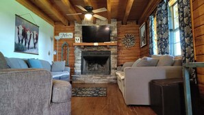 A cozy living room with a stone fireplace, wooden ceiling beams, and a television mounted above it. - 37 acres, fishing, hunting, golf course nearby, rustic feel, hiking, near town. (Pocahontas)