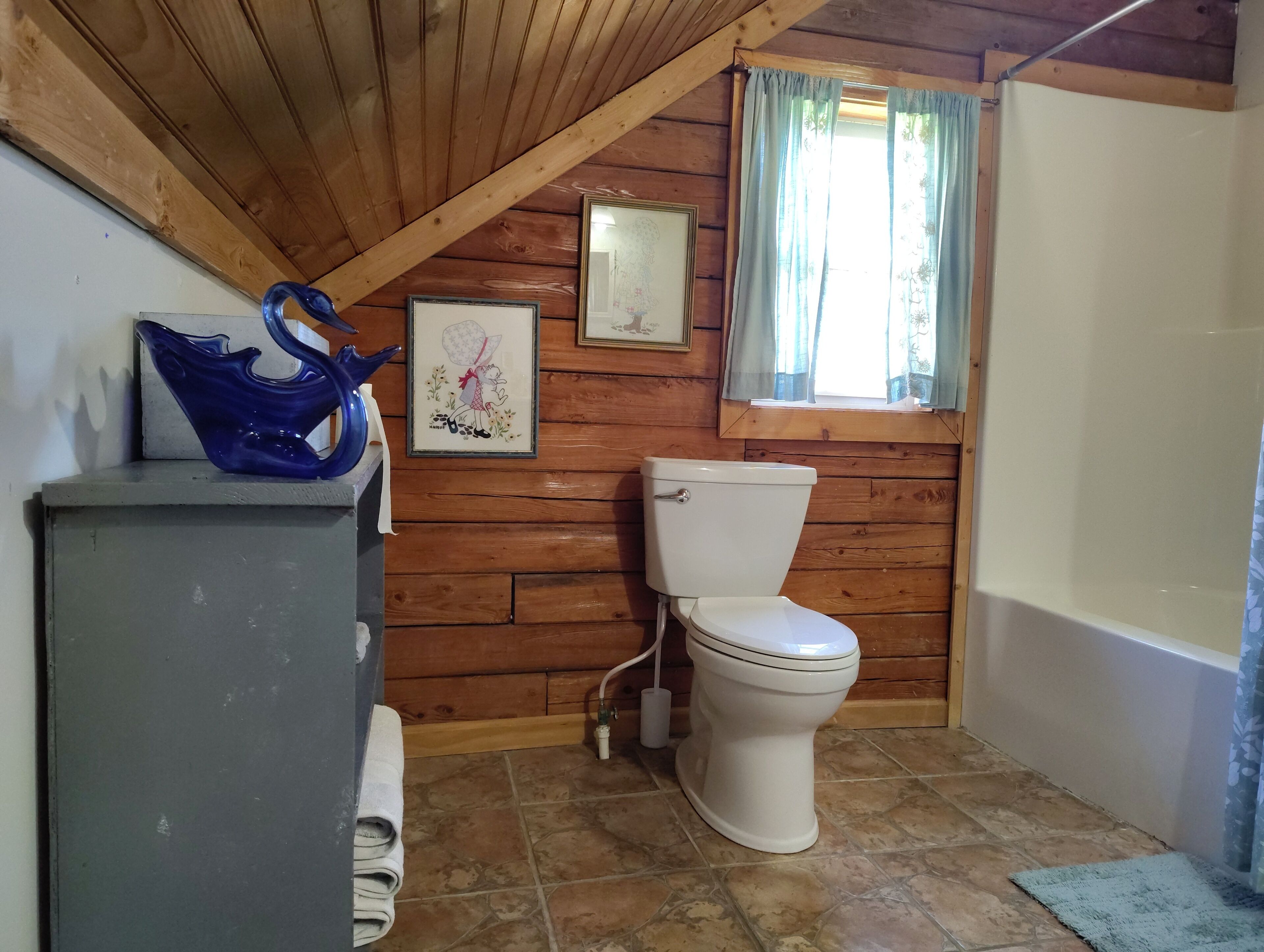 A wooden-paneled bathroom with a toilet, a framed picture, and a blue glass vase on a cabinet.